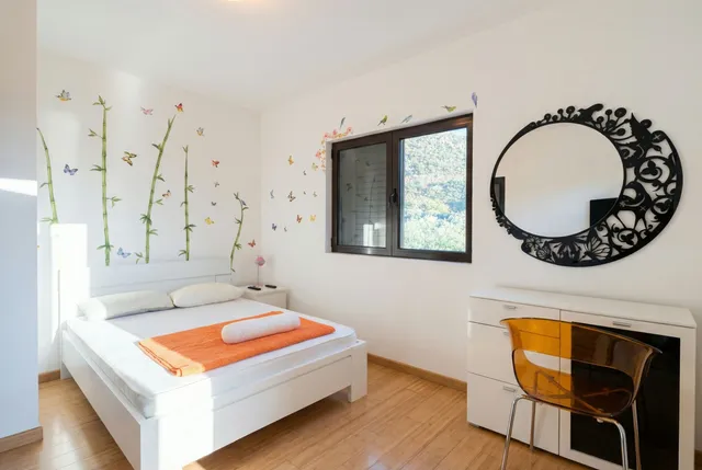 Светлая спальня в квартире в Бечичи, Черногория: двуспальная кровать, дизайнерское зеркало и вид на горы из окна.