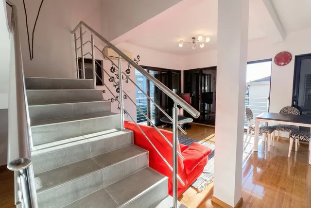 Интерьер двухуровневой квартиры в Бечичи, Черногория: лестница с перилами, красный диван и обеденная зона у окна.