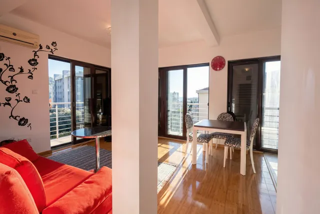 Светлая гостиная с красным диваном и обеденной зоной в квартире в Бечичи, Черногория, панорамные окна с видом на город.